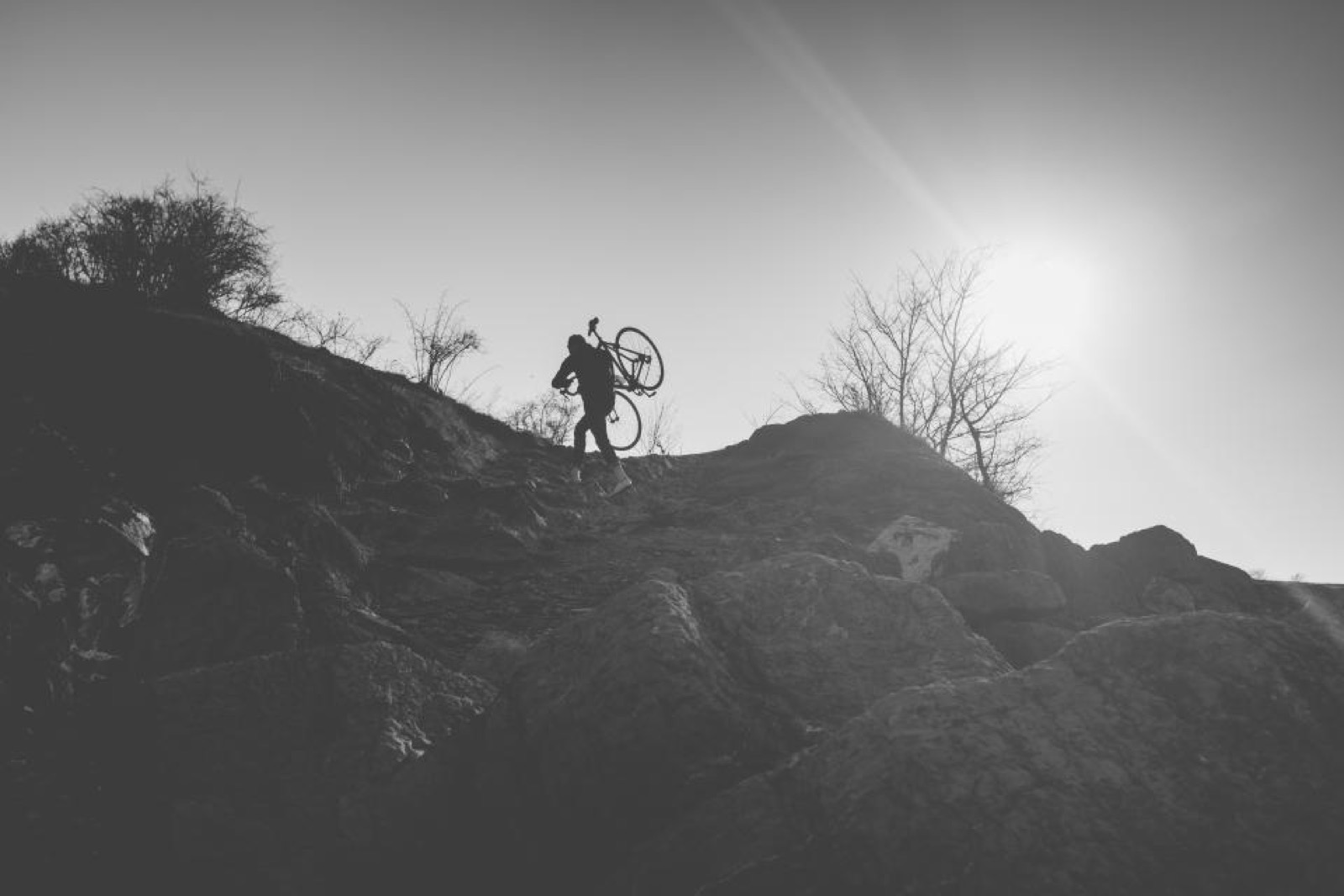 A rider silhouetted against the sky carrying a bike up a rocky ridge